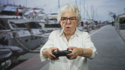 Senior woman holding gamepad with both hands pointed forward on a marina pier, smiling and reacting as she plays  playful joy. © Krakenimages.com
