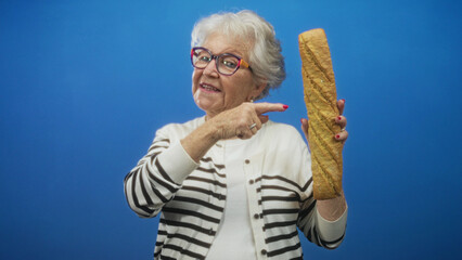Senior woman points with hand at a long baguette while holding it upright against a blue studio backdrop  playful baking nostalgia. © Krakenimages.com
