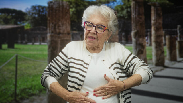Senior woman clutching chest and abdomen, eyes closed, wearing striped cardigan and glasses, standing among ancient columns at a building site; pain concern vulnerability.