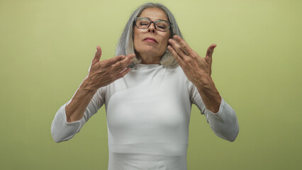 Naklejka premium Middle aged grey haired woman with hands on chest and glasses in studio green backdrop; self care serenity.
