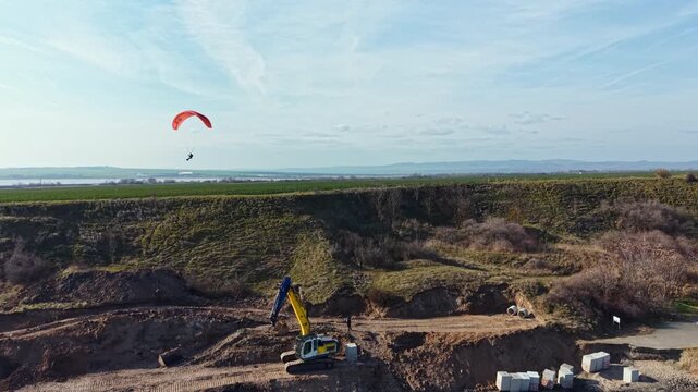 A person is paragliding over a construction site located in a rural area. The scene shows machinery and a vast open field under a clear sky.