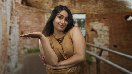 Fototapeta premium Young woman shrugging with palm up and bare shoulders amid roman ruins building, leaning on metal railing and glancing aside; curiosity contemplative.