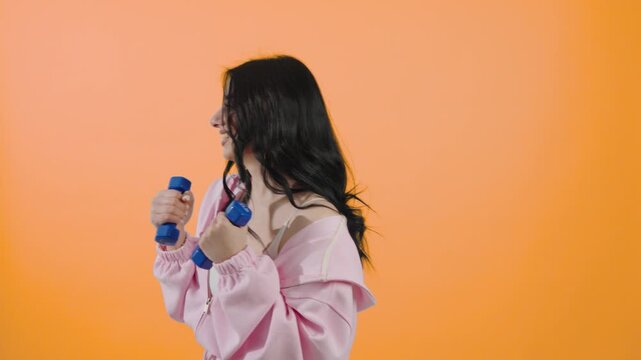 Young woman training with blue dumbbells on orange background