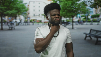 Fototapeta premium Man clutching throat in pain on a city street, eyes closed and wearing white t shirt and bead necklace; distress.