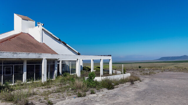 Aerial view of an abandoned white building with a red tiled roof and overgrown vegetation overlooking a vast open landscape under a clear blue sky in iNtabazwe, Free State, South Africa.