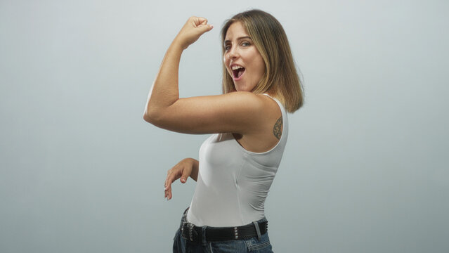 Woman flexes bare upper arm showing bicep in studio wearing white tank top and jeans, playful pout and confident pose; confidence strength empowerment.