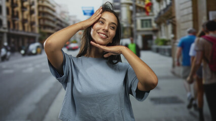 Fototapeta premium Woman posing with hands framing face, smiling in a casual tshirt on a busy street in city center; playful confidence.