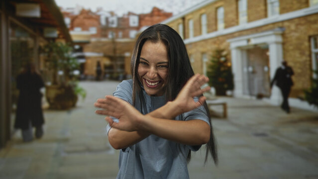 Young hispanic woman in light blue shirt laughing with bare hands reaching forward and eyes closed on busy street with brick building facades; joy celebration.