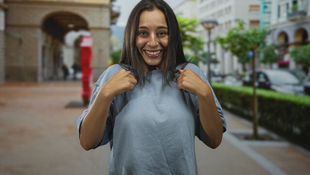 Woman pulls up shirt to reveal stomach while smiling and raising both arms on street with arched building and parked car; playful.
