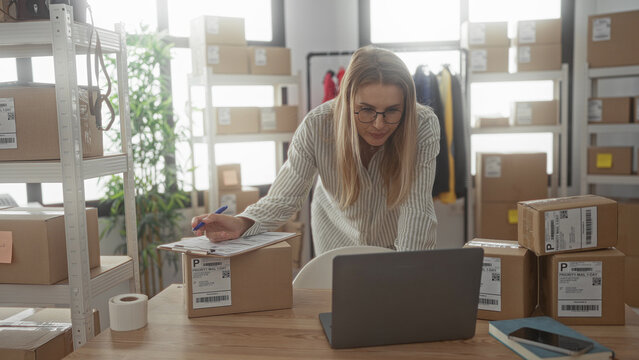 Woman leaning over laptop and signing shipping label on cardboard box amid stacked parcels and shelves in studio; small business focused.