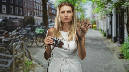 Obraz premium Woman holding a vintage camera with palm raised in a stop gesture on a city street, white dress visible and gaze fixed; annoyance travel.