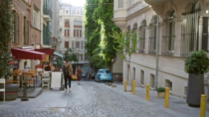 Fototapeta premium Narrow cobblestone street with soft bokeh and defocused shallow focus on storefronts, trees and parked car, outdoor; background copyspace calm.