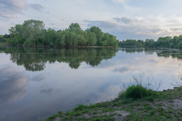 Fototapeta premium a calm river reflecting lush green trees and a cloudy sunset sky.