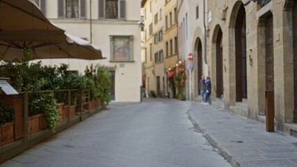Fototapeta premium Quiet narrow city street in soft defocused bokeh showing blurred stone facades, cafe planters and umbrella, shallow focus outdoor background; background backplate copyspace calm.
