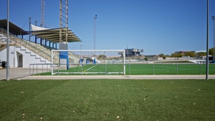 Obraz premium Soccer pitch foreground softly blurred with shallow focus on turf and defocused goal and stadium stands visible outdoor; backdrop copyspace backplate template.
