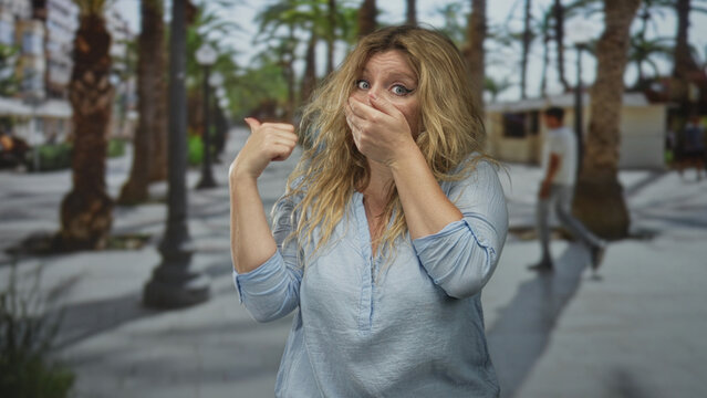 Woman covering mouth with hand and pointing thumb over shoulder on a street lined with palm trees; shock reaction.