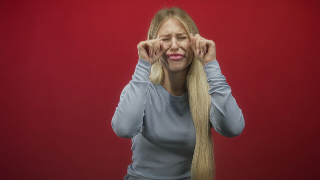 Woman pinches her lips with fingers and pinches cheeks, eyes closed while wearing a light blue sweater against a studio red backdrop; playful gesture.