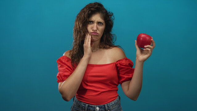 Young hispanic woman holding red apple with hand on cheek in teal studio, wearing off shoulder red top and denim jeans; disgust toothache.