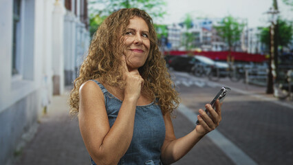 Fototapeta premium Woman points index finger to smartphone on street, tapping screen and smiling while checking messages; lighthearted joy.