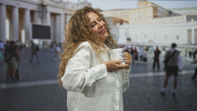 Middle aged woman holds a mug reading i am the boss and cradles it to her chest, smiling in front of a large building; quiet confidence.
