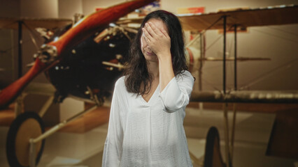 Woman covers face with hand and makes expressive hand gestures beside a vintage biplane propeller inside building  frustration reflection. © Krakenimages.com