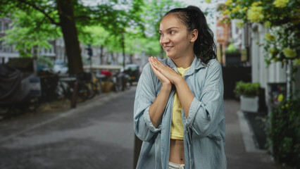 Fototapeta premium Young woman clapping hands and showing bare midriff on a tree lined street with bikes and planters; joyful.