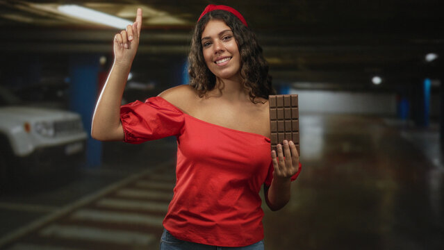 Woman points finger up and holds chocolate bar in an underground parking garage while smiling and wearing a red off shoulder top and headband; happiness.