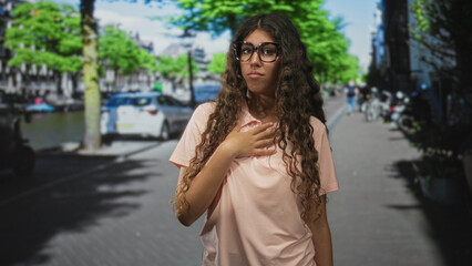 Fototapeta premium Woman touching chest with hand and slight grimace, wearing glasses and pink shirt on street; embarrassment.