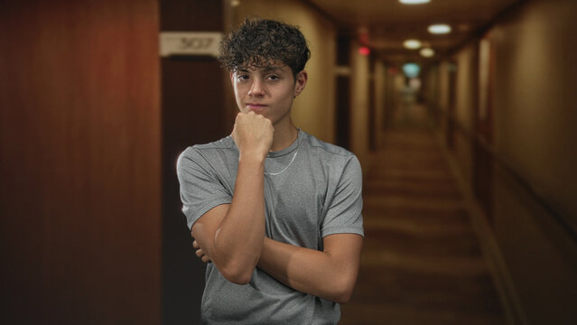 Teenage boy with hand on chin and crossed arms, wearing silver chain, standing by door 307 in hotel building corridor; thoughtful reflection.