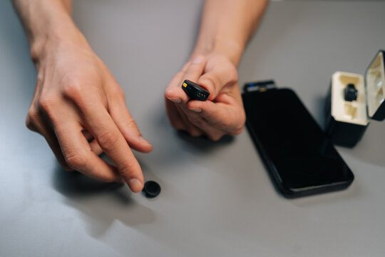 Close-up of hands holding compact wireless lavalier transmitter and magnetic plate next to smartphone receiver on gray surface, preparing to attach mic for vlogging and portable audio recording.