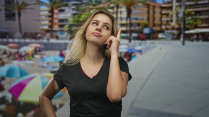 Fototapeta premium Young blonde woman points finger to temple while standing near a beachfront building in a vneck tshirt with hand on hip; thoughtful curiosity.