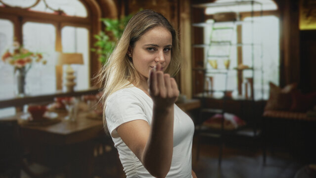 Young blonde woman in white t shirt beckons with fingers in living room, looking directly at camera with a serious expression; challenge.