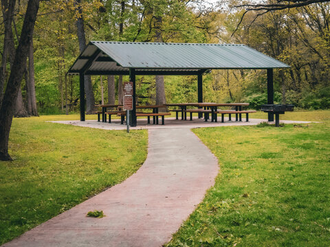 Empty outdoor picnic shelter with tables and a charcoal grill at the end of a paved path in a lush green forest preserve during the spring season