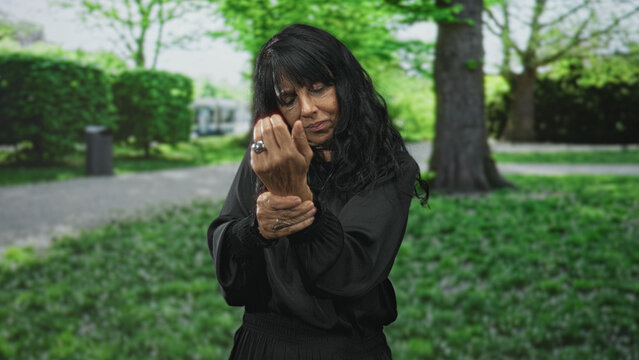 Woman holding and rubbing wrist, wearing rings and a black blouse while looking down on street path among trees; pain resilience.