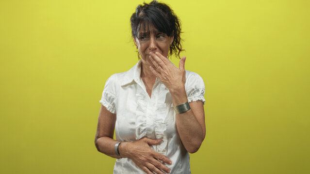 Woman with hand to mouth and other hand pressed on stomach, wearing white blouse and bracelets against yellow backdrop in studio, grimace showing queasy reaction; nausea discomfort.