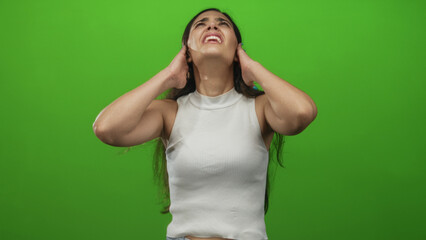 Fototapeta premium Woman with long dark hair in white tank top grimacing while covering ears in green studio; stress.