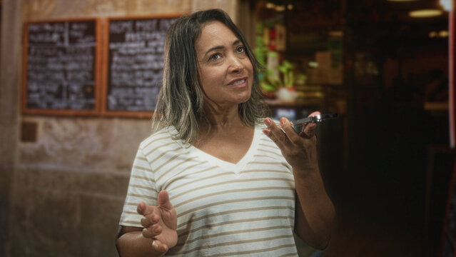 Hispanic woman holding smartphone and gesturing while speaking into phone in a building cafe interior with chalk menu and counter; frustration.