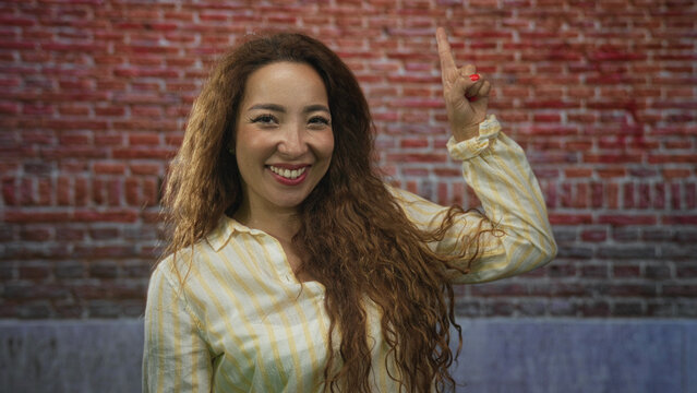 Woman in yellow striped shirt points finger upward at brick building showing open smile and long hair; joyful confidence.