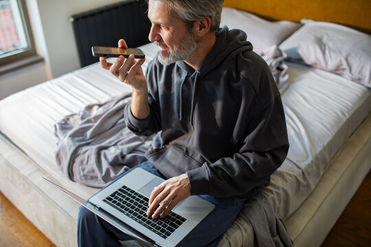 Man using voice command on phone while working on laptop in bedroom