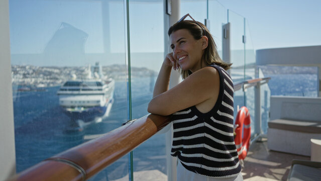Woman leaning on cruise deck rail with bare arms resting on wooden handrail, smiling and looking at a distant ship; serenity.