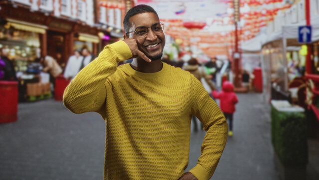 Young african man with hand to ear making a phone call gesture on a crowded street wearing yellow sweater and glasses; connection joy.