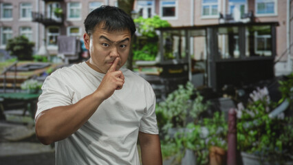 Fototapeta premium Man in white t shirt making finger to lips for silence on an urban street with brick facades and potted plants; secrecy.