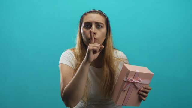 Woman holding a pink present and pressing finger to lips in studio, leaning forward with worried glance and tense expression; secret silence.