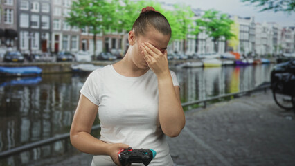 Woman holding gamepad with hand covering face on street canal walkway in front of buildings, wearing white tshirt  frustration defeat. © Krakenimages.com