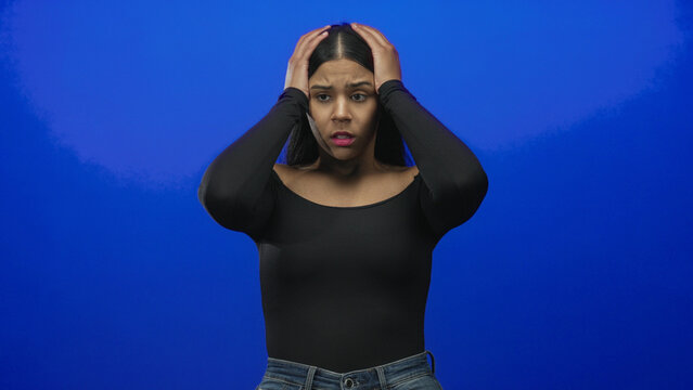 Young latin woman grasping head with both hands in vibrant blue studio wearing black offshoulder top and denim jeans; anxiety.