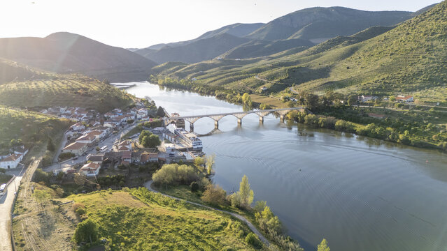 Aerial view of Barca de Alva village and the bridge over the Douro River, surrounded by terraced vineyards and green hills in Figueira de Castelo Rodrigo, Distrito da Guarda, Portugal.