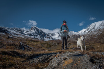 Female hiker with trekking poles and white dog walking in snowy mountains. Active woman traveler with husky on trail, autumn alpine landscape, wilderness adventure and pet travel concept © ANDREY