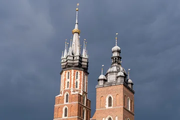 The asymmetric towers of St. Mary's Basilica in Krakow. A detailed view of the Gothic brick church's spires against a dark, stormy sky in the Main Market Square, Poland. © Pat dream