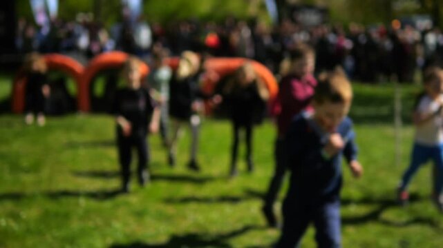 Defocused shot of energetic children participating in a kids' marathon, running across a sunny green field with a crowd and orange inflatable obstacles cheering them on in the background
