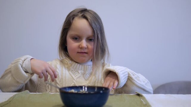 Fussy little girl sitting at a table, making faces and showing disgust while refusing to eat her morning cereal. She plays with the spoon and pushes the bowl away in a display of pickiness